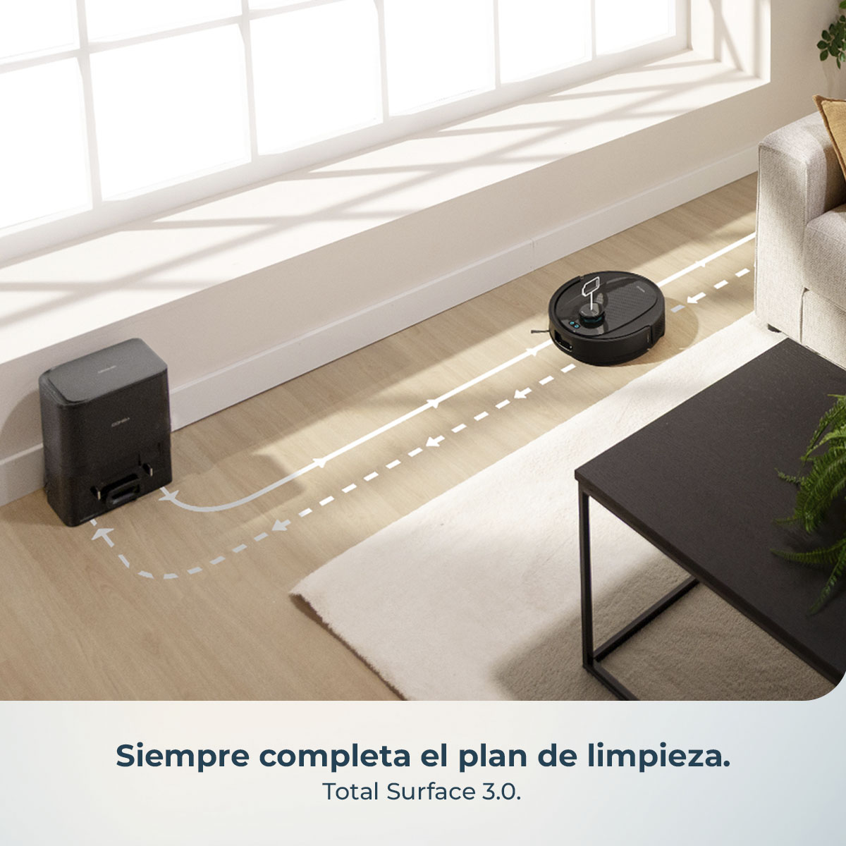 Una aspiradora robot y su estación de carga están en una habitación. Se muestra una ruta de limpieza. También hay un sofá, una alfombra y una mesa.
