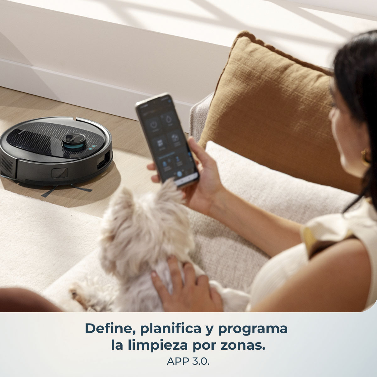 Una mujer usa un teléfono para controlar una aspiradora robótica. Un perro blanco pequeño está en primer plano. Una almohada marrón en el fondo.
