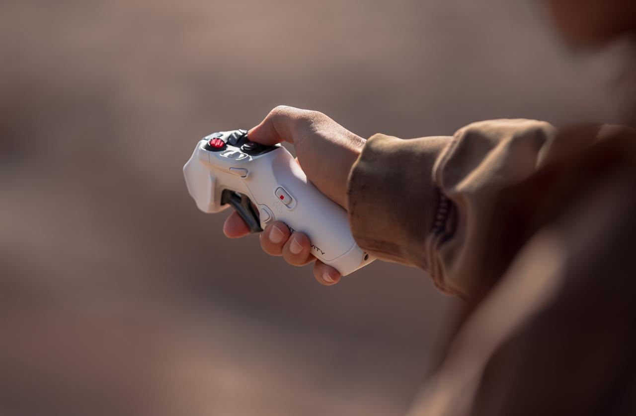 Eine Hand hält eine weiße Drohnen-Fernbedienung. Der Controller hat schwarze Tasten und rote Akzente. Der Hintergrund ist braun verschwommen.