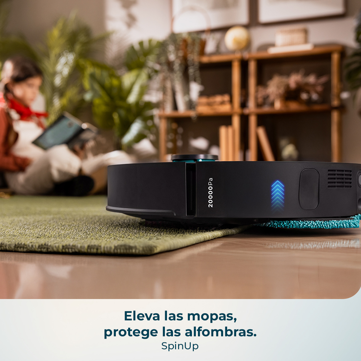 Una aspiradora robot negra sobre una alfombra verde. Una persona lee en el fondo. También se ven estanterías de madera y plantas.