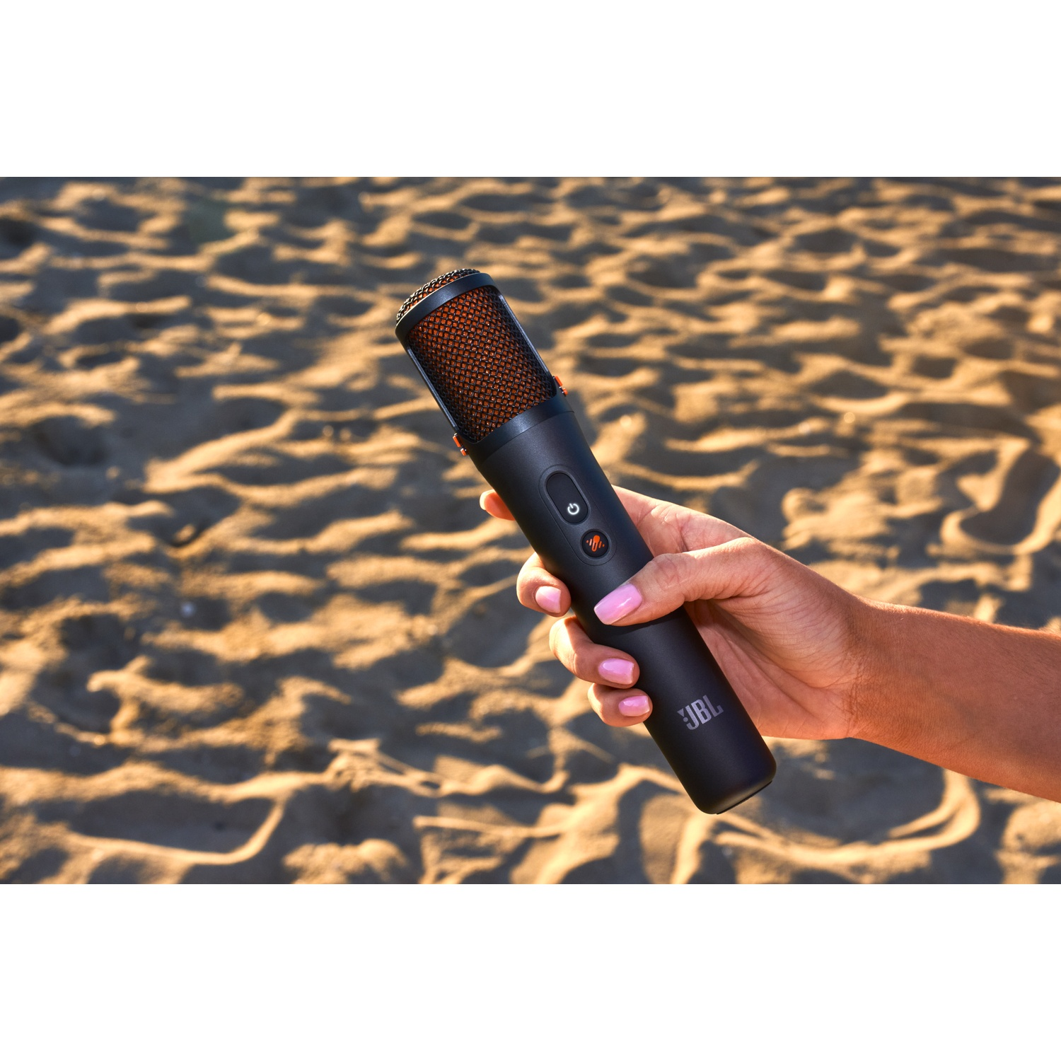 Een hand houdt een zwarte JBL-microfoon vast op een zandstrand. De microfoon heeft een oranje mesh bovenkant.