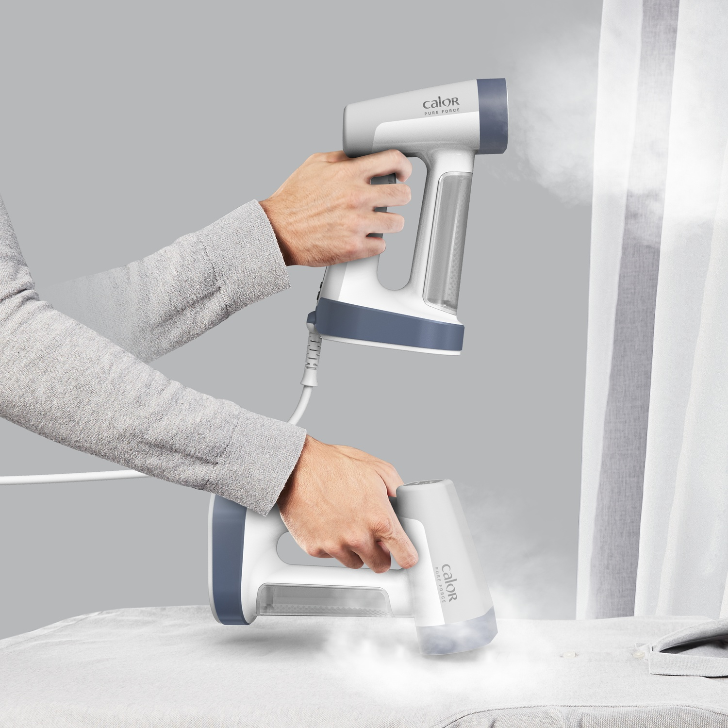 Hands holding a white and blue steam iron. One iron is steaming fabric, another is held up. A gray curtain is in the background.