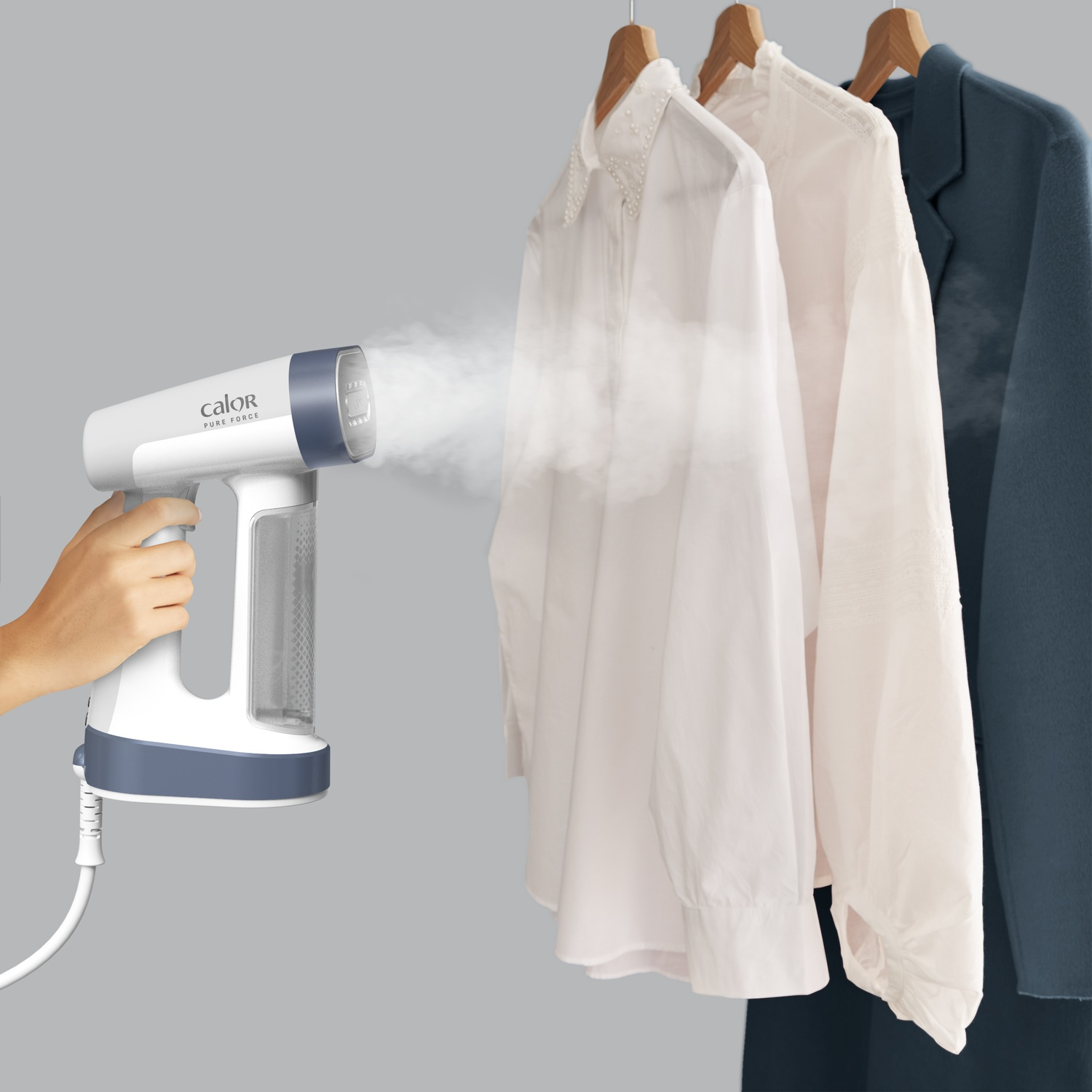 A hand holds a white and blue garment steamer, steaming white shirts and a blue blazer hanging on wooden hangers.