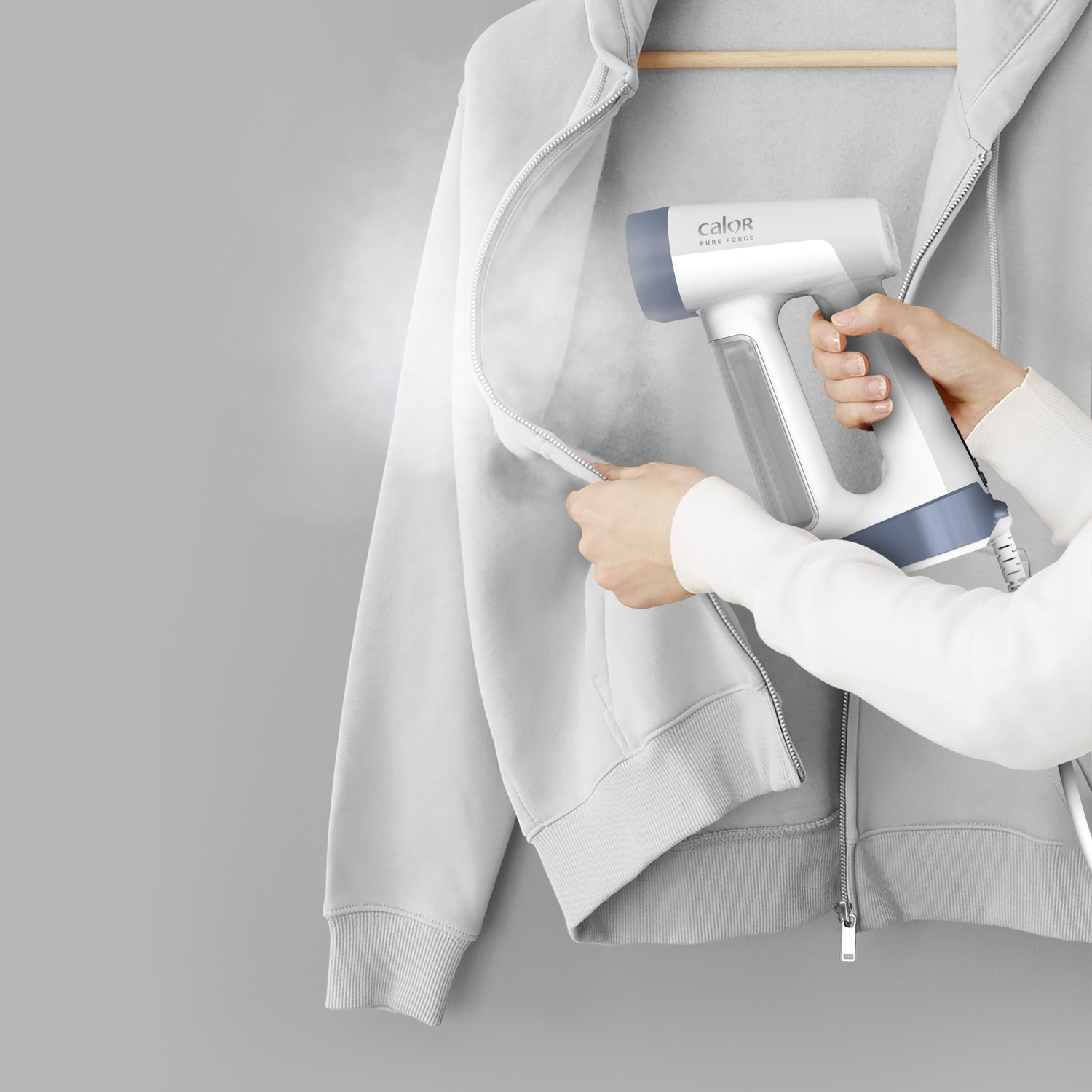 A person uses a steamer on a gray hoodie. The steamer is white and gray. The background is a gray wall.