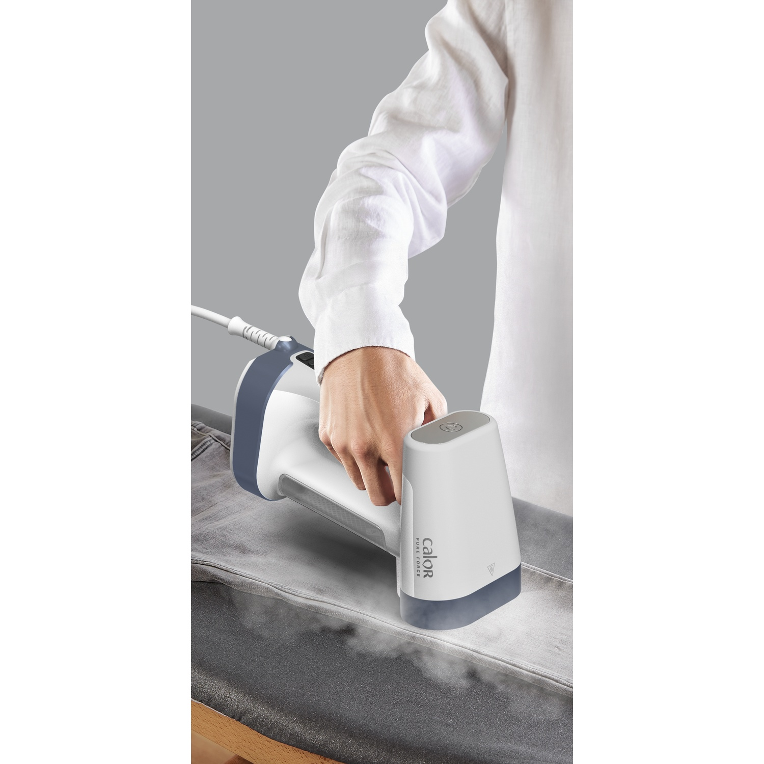 A person ironing grey denim with a white and blue garment steamer. Steam rises from the fabric. The background is grey.