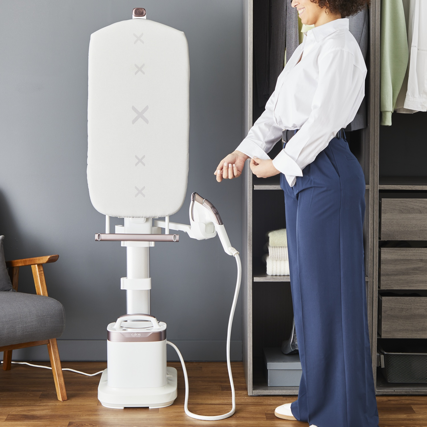 Woman adjusts sleeve in front of closet. A garment steamer is on the left. White shirt, blue pants.