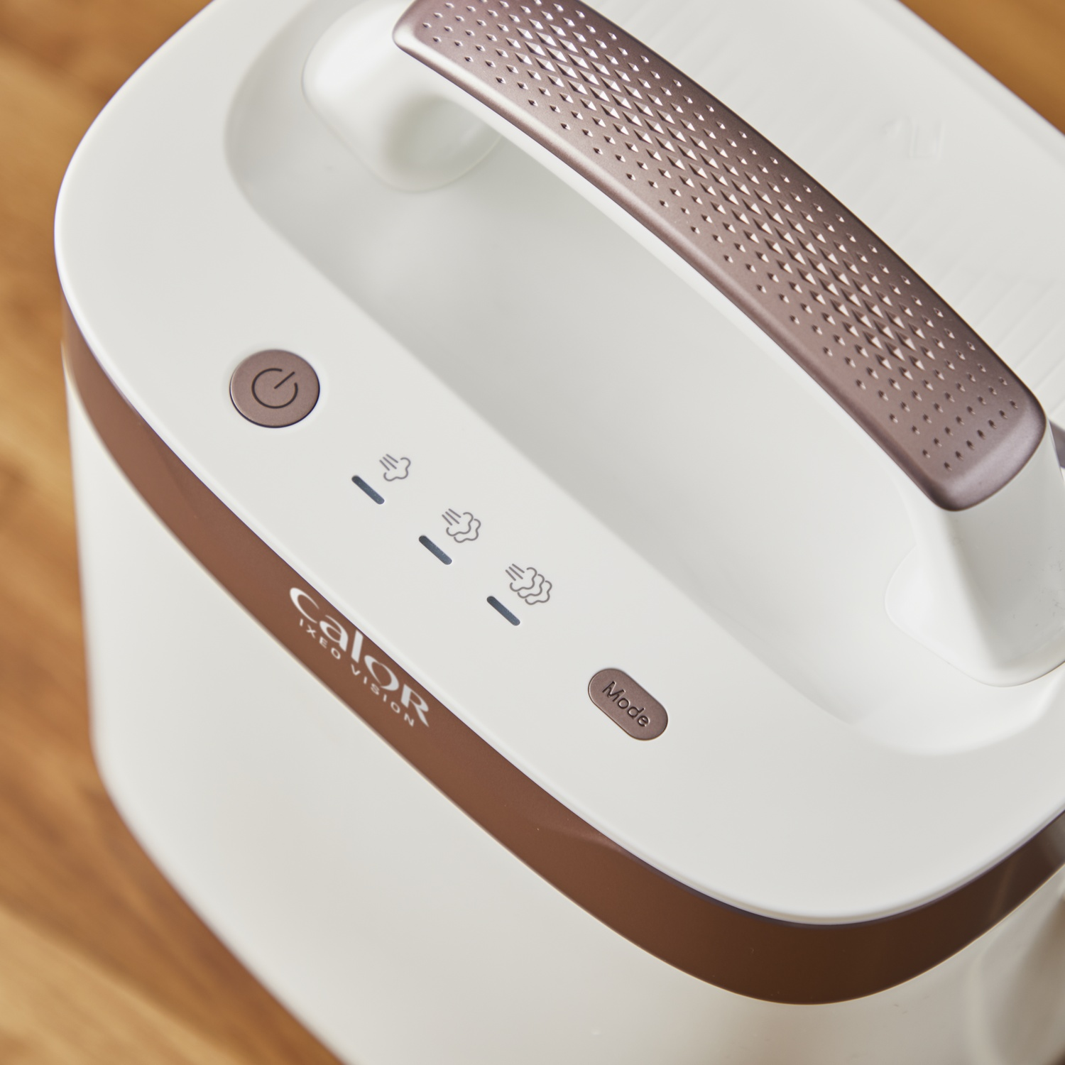 Close-up of a white and brown steam iron. It has a handle and control buttons for power and steam settings. Wooden surface in the background.