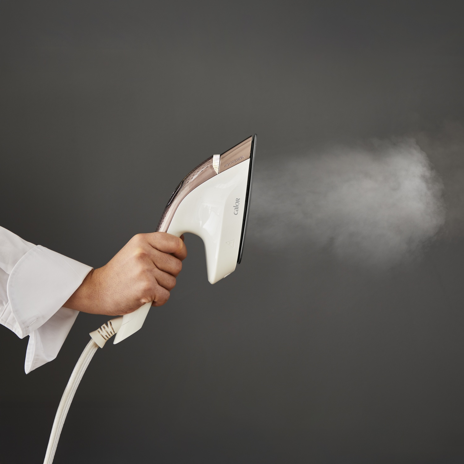 A hand holds a white and gold iron, releasing steam against a gray background. A white shirt sleeve is visible.