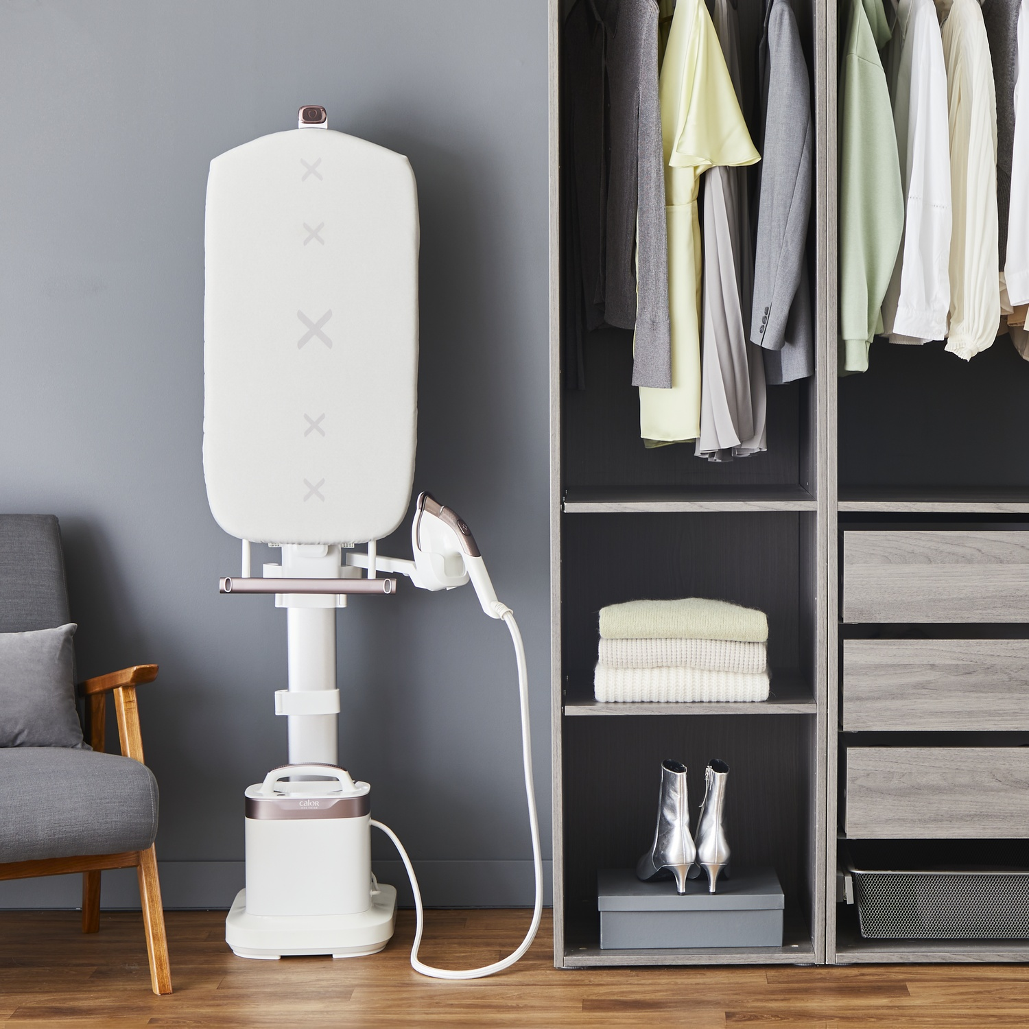 A white and brown clothes steamer stands next to a wooden wardrobe filled with clothes. A grey armchair is on the left.