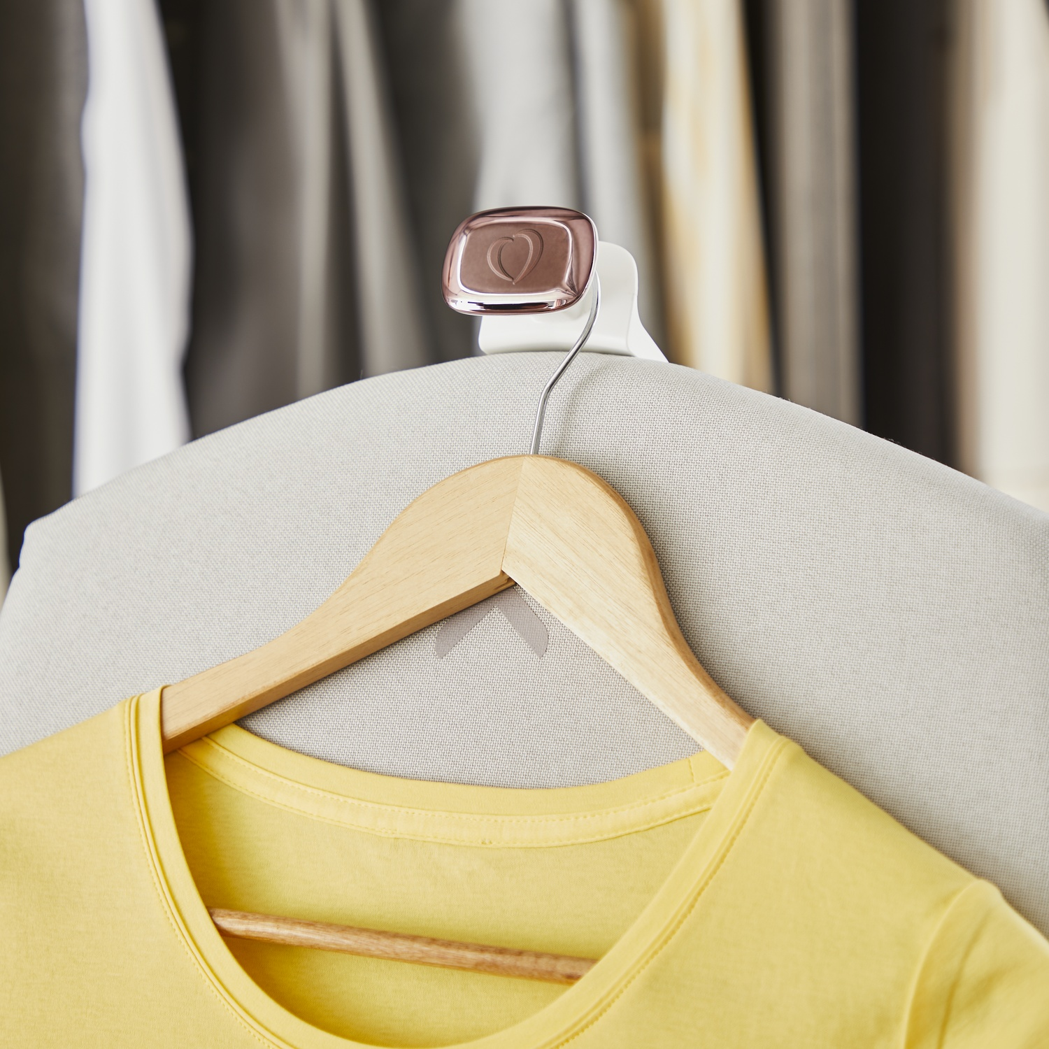 A yellow t-shirt hangs on a wooden hanger. A heart-shaped clip is attached to the hanger. Clothes are visible in the background.