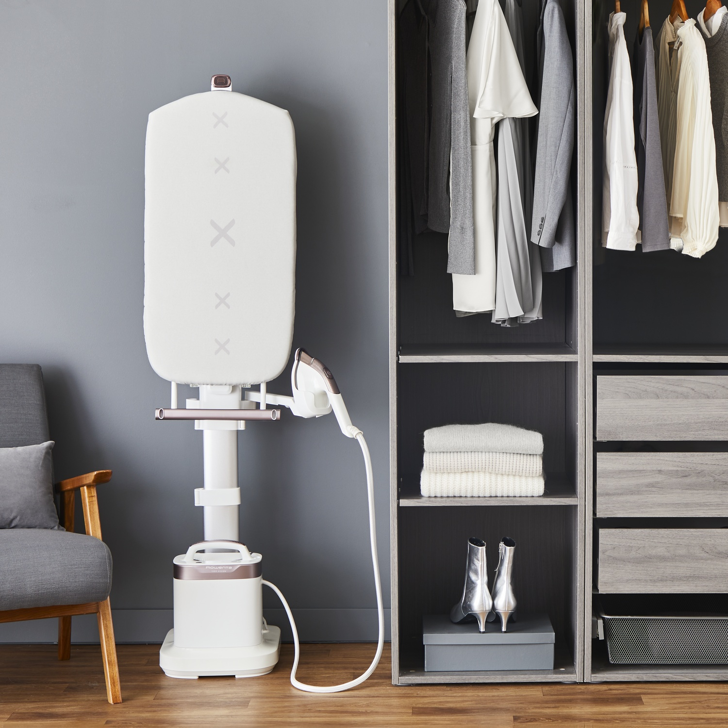 A white garment steamer stands next to a gray armchair and a closet filled with clothes. Silver boots sit on a shelf.