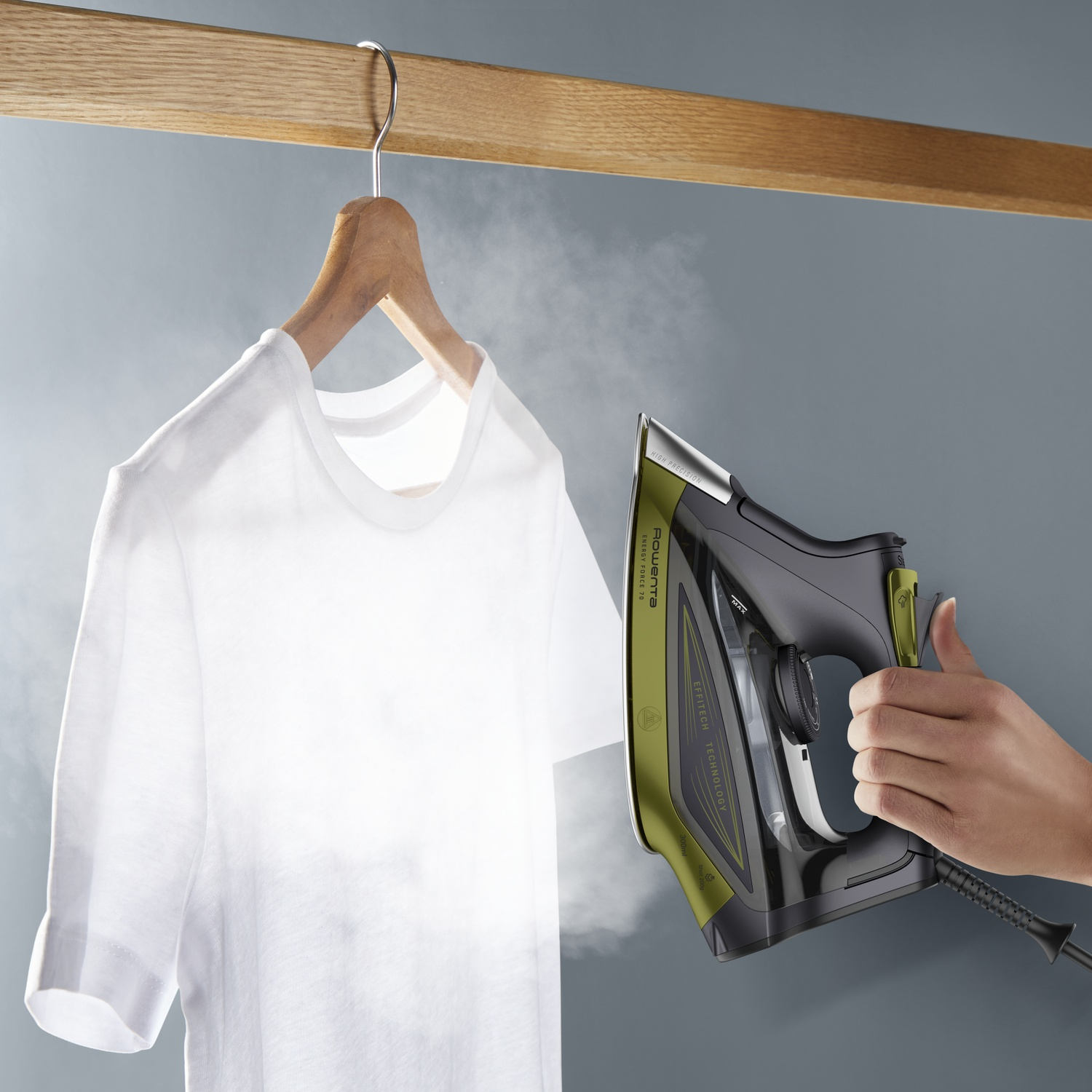 Un t-shirt blanc est suspendu à un cintre en bois. Une main tient un fer à repasser noir et vert, vaporisant la chemise. Fond gris.