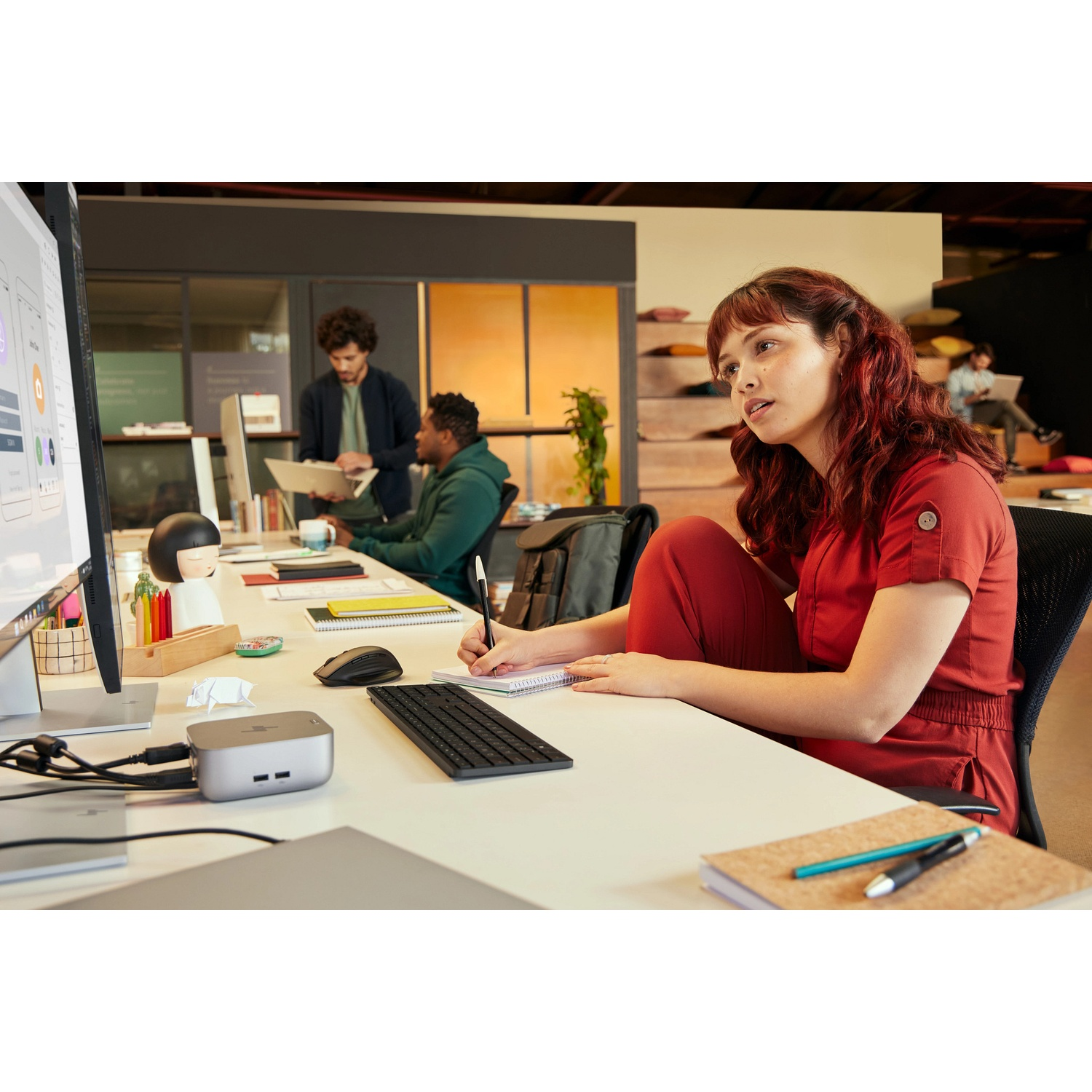 Een vrouw in het rood schrijft aan een bureau. Computer, toetsenbord en andere werknemers zijn op de achtergrond. Notitieboekje en pen aanwezig.