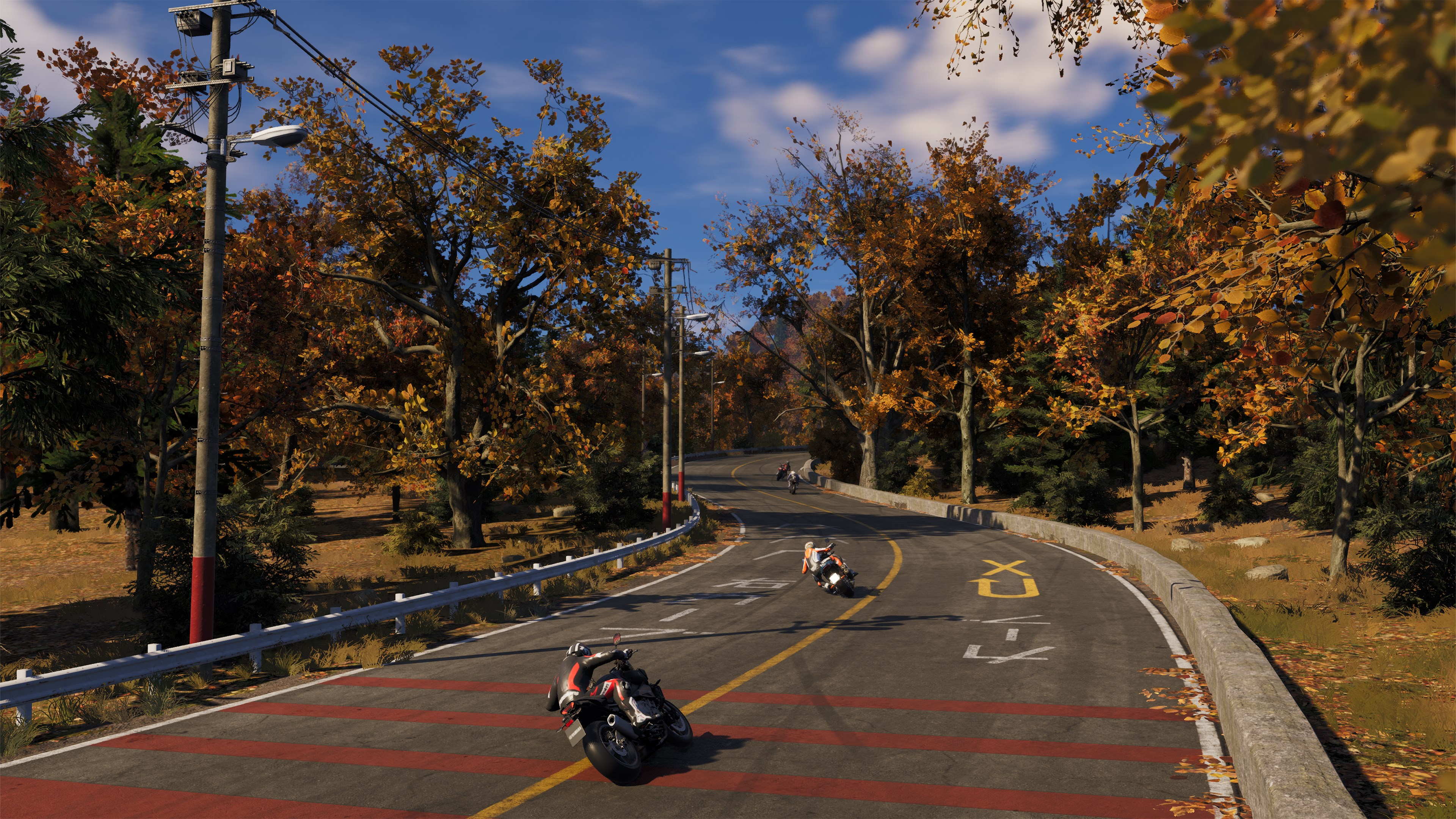Des motocyclistes roulent sur une route sinueuse à travers une forêt avec un feuillage d'automne. Le ciel bleu est visible. Plusieurs motos sont en mouvement.