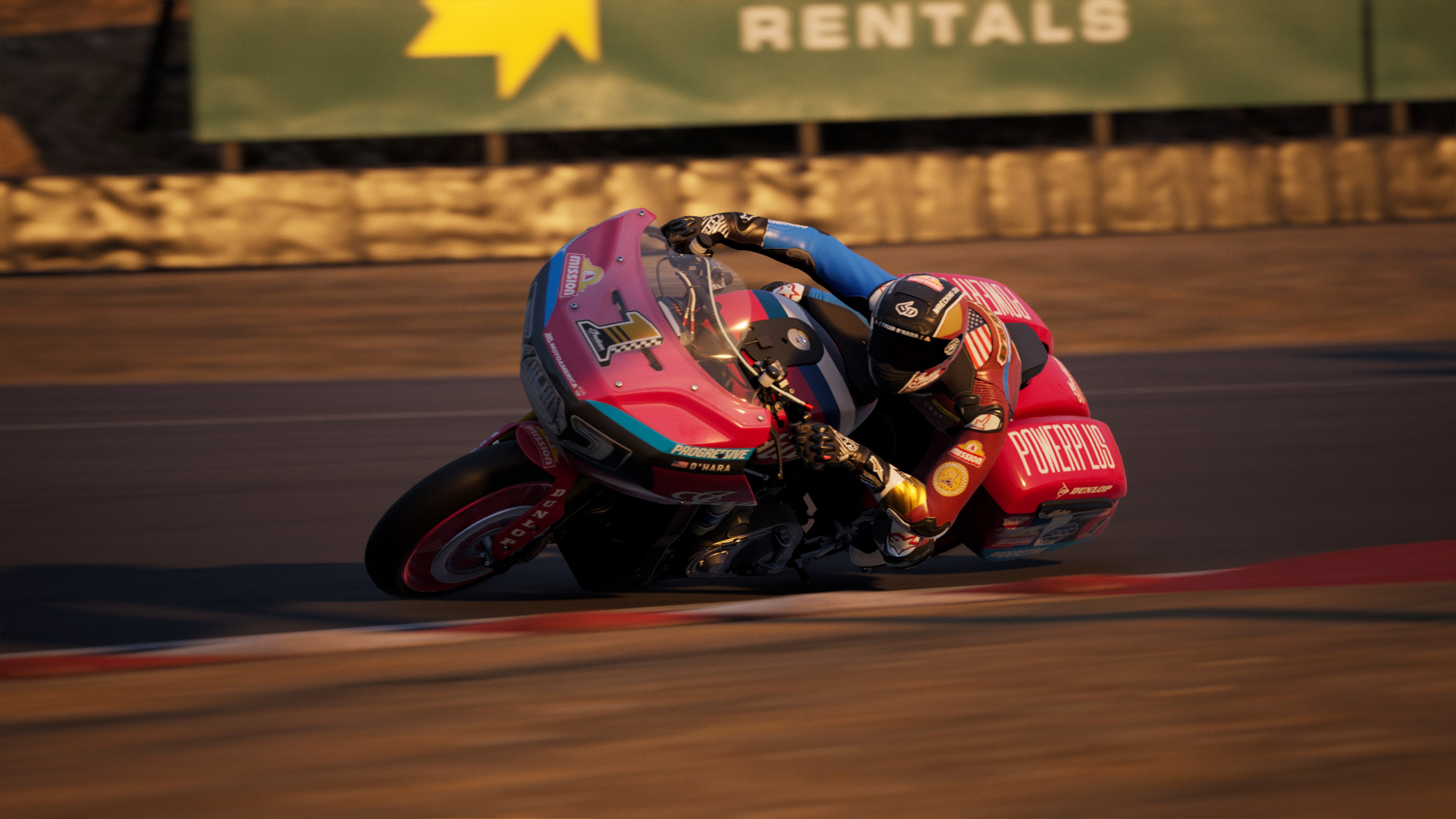 Un motocycliste se penche dans un virage sur une moto rose. La scène se déroule sur un circuit. Panneau 'RENTALS'.
