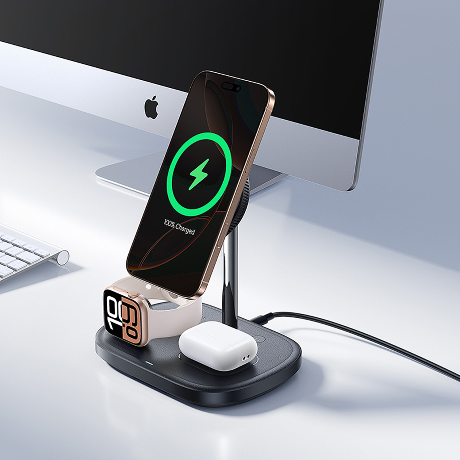 Una estación de carga con teléfono, reloj y auriculares. El teléfono muestra un icono de carga. Un ordenador Apple en el fondo.