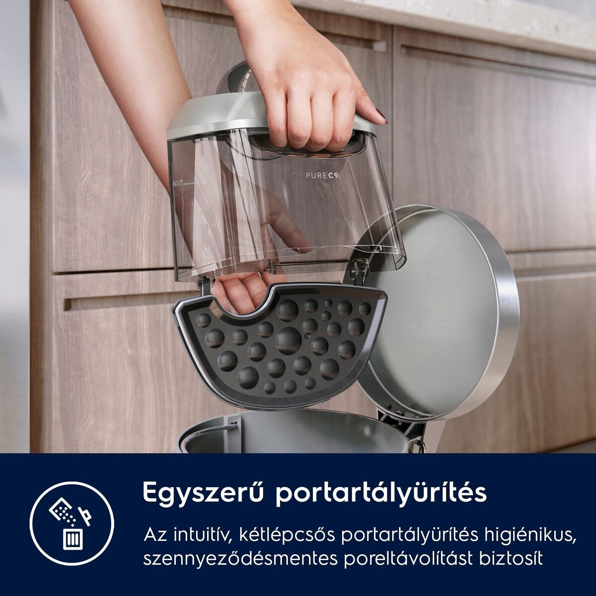 Egy személy egy átlátszó tartályt tart szürke fedéllel, egy porszívó részét, egy fa szekrény közelében.