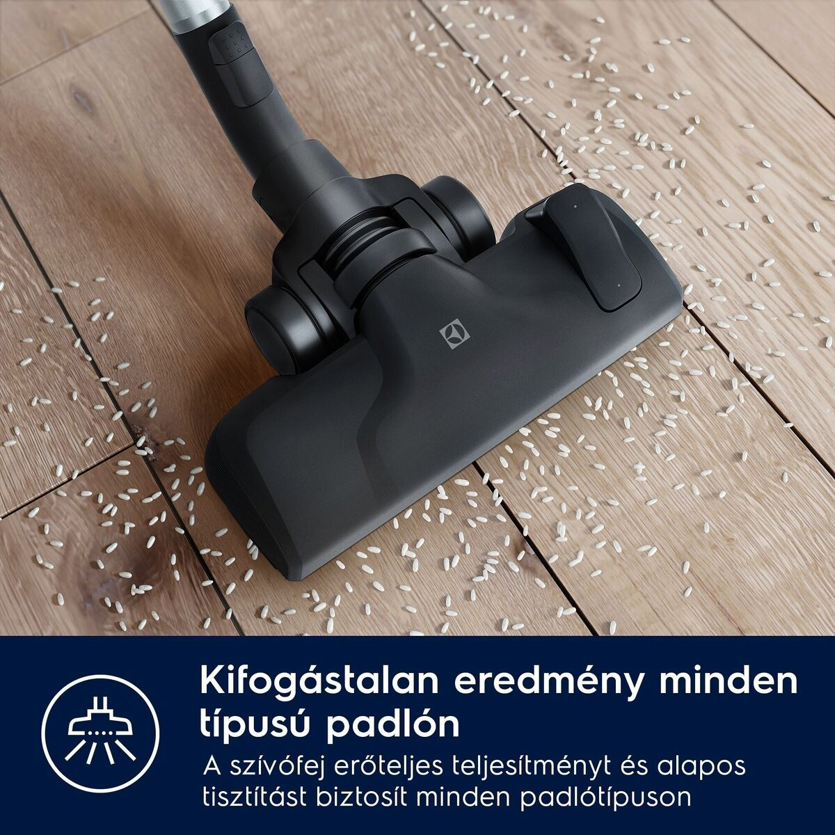 Fekete porszívófej egy fa padlón, szétszórt fehér rizzsel. A porszívó használatban van.