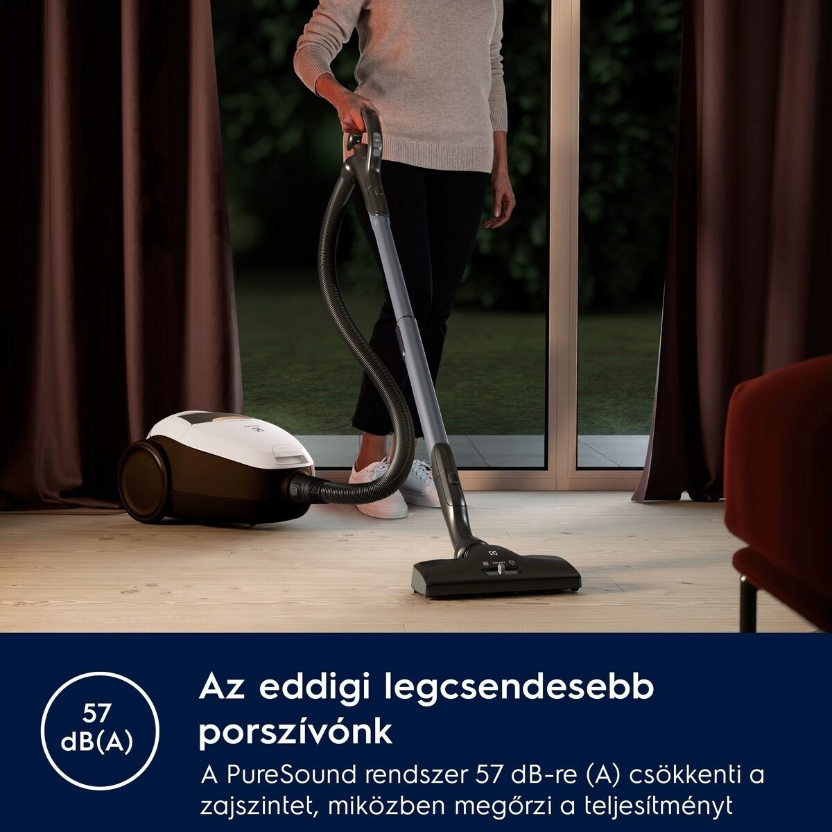 Egy nő porszívót tart. A porszívó a padlón van. A háttérben ablak, függönyök és egy fotel látható.