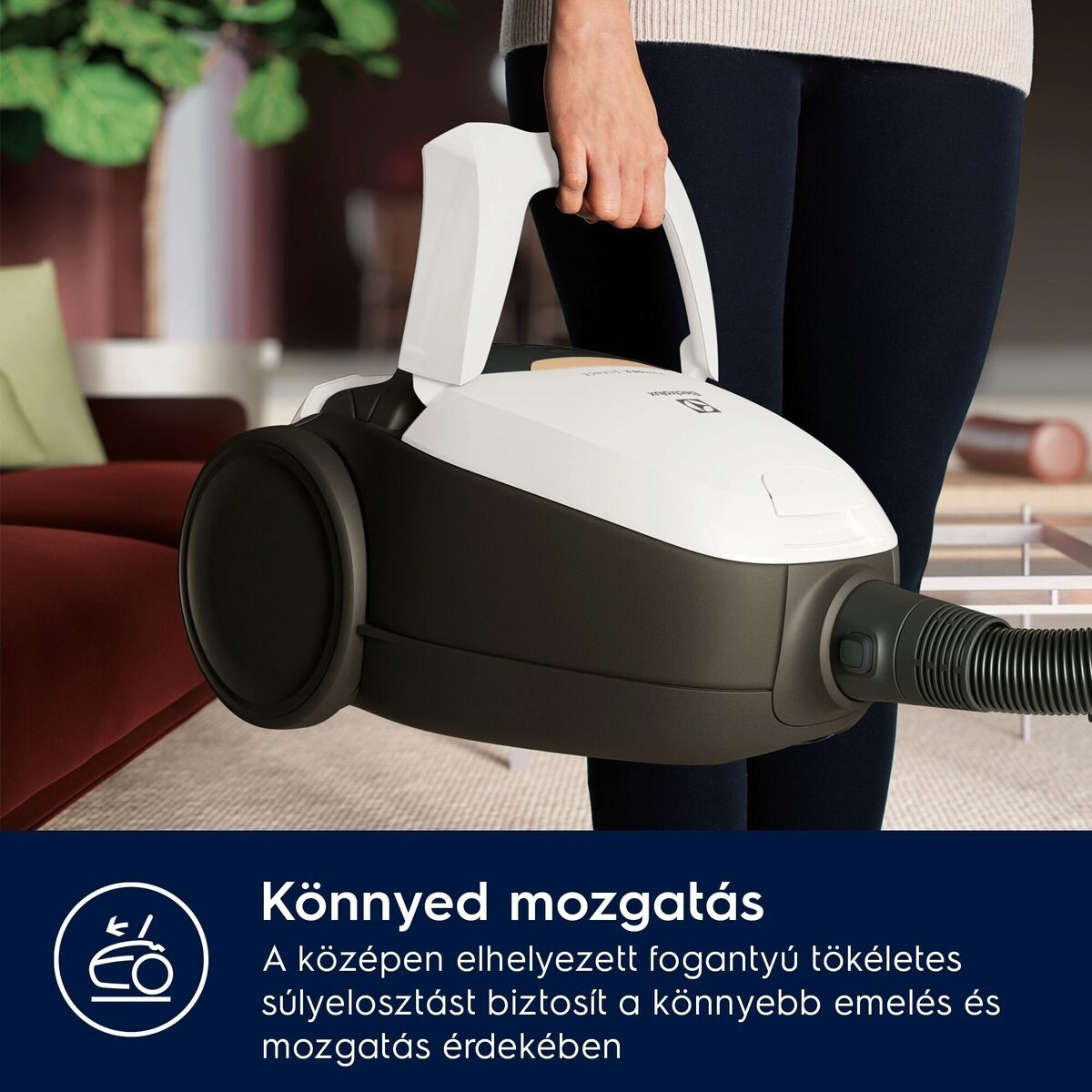 Egy személy egy fehér és sötétszürke porszívót tart. A porszívón van egy fogantyú. A háttérben egy szoba és szöveg látható.