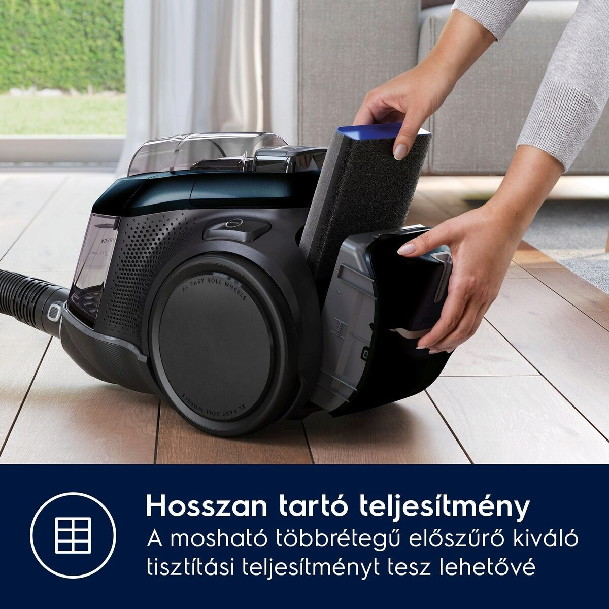Egy nő fekete szűrőt helyez egy fekete porszívóba. A porszívó egy fa padlón van, egy ablak közelében.