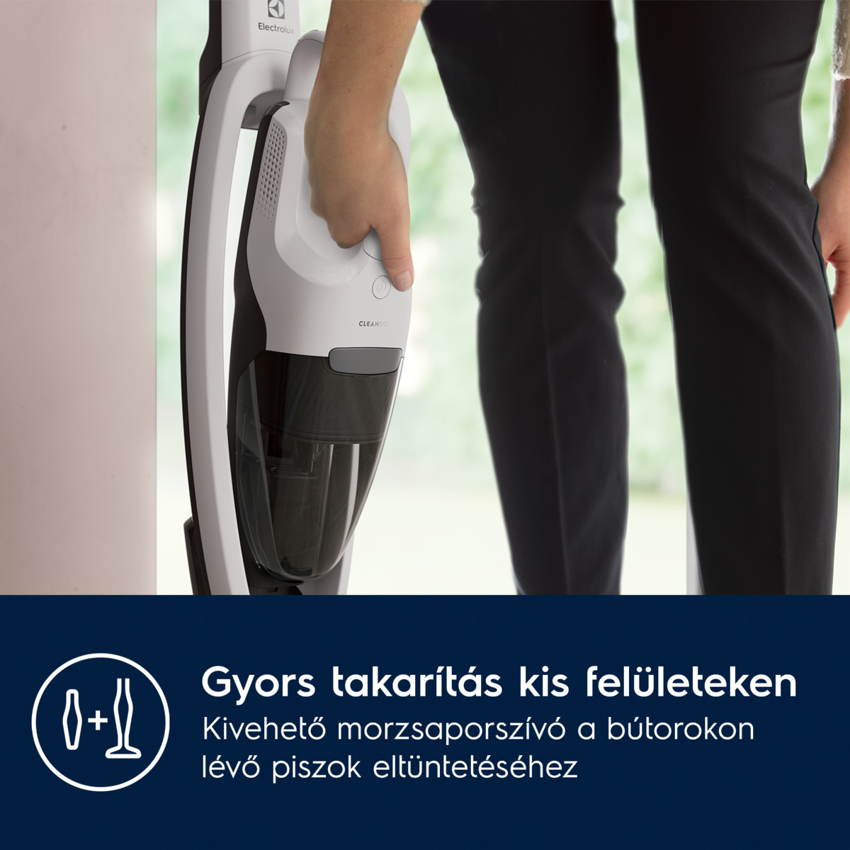 Egy személy egy fehér és fekete porszívót tart. A porszívó a fal közelében van. A személy fekete nadrágot visel. Az edény átlátszó.