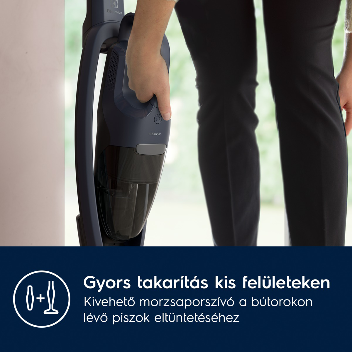 Egy személy egy sötétkék vezeték nélküli porszívót tart. A porszívó fogantyúját a személy keze szorítja. Fekete nadrág látható.