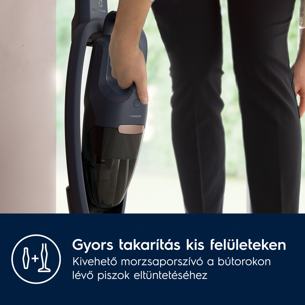 Egy személy egy sötétkék kézi porszívót tart. A porszívón van egy fogantyú és egy portartály. A személy fekete nadrágot visel.