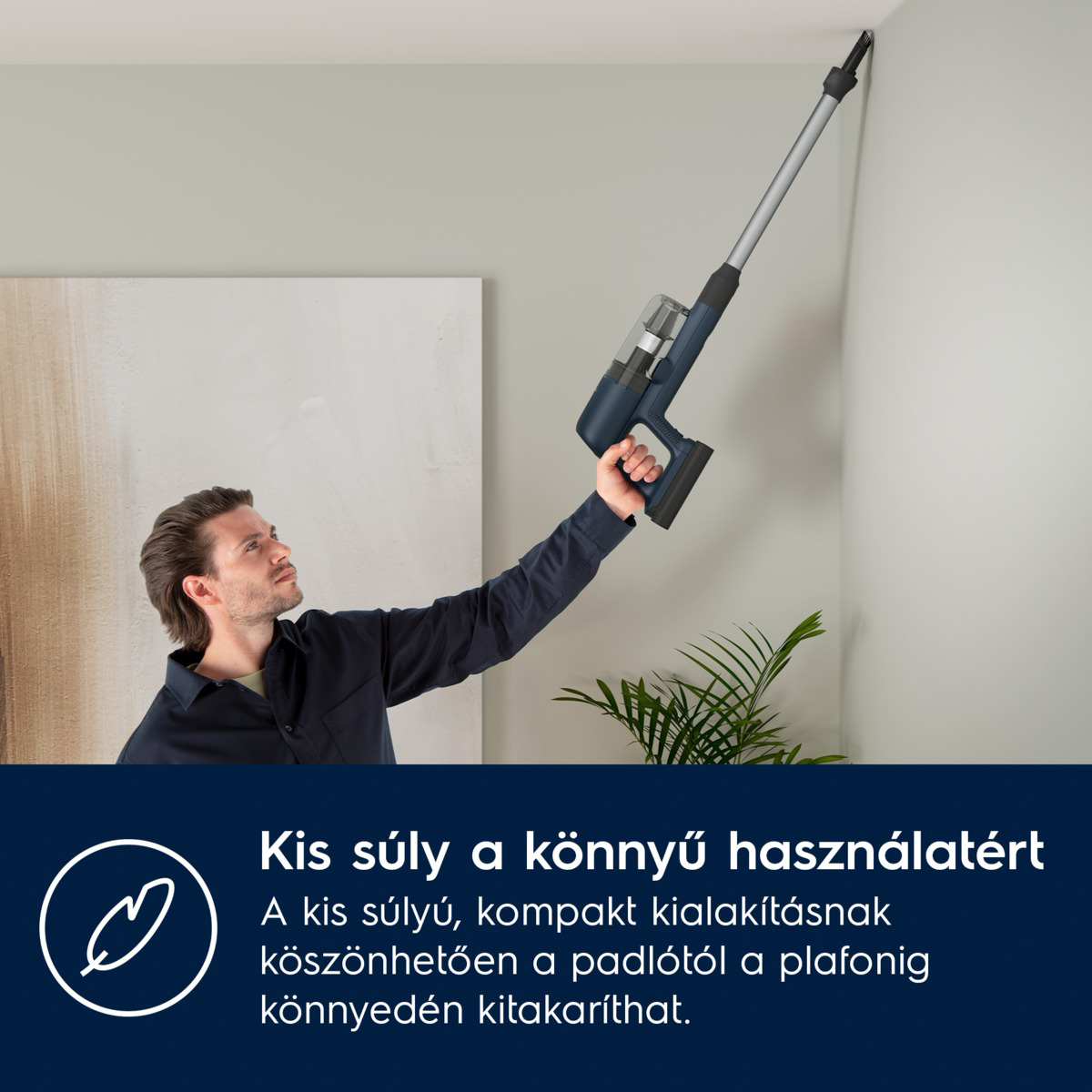 Egy férfi egy kék porszívót tart a kezében, és a mennyezetet tisztítja. A háttérben festmény, növény és fehér fal látható.