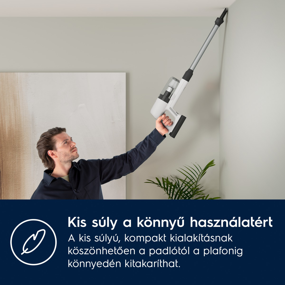 Egy férfi egy fehér-szürke porszívóval takarítja a mennyezetet. Felfelé néz. A háttérben egy növény látható.