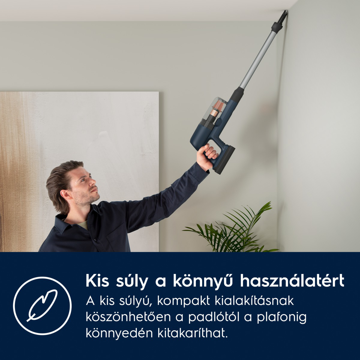 Egy férfi egy kék porszívót tart egy sarok felé. Egy növény és szöveg van az előtérben. A háttér egy fehér fal.