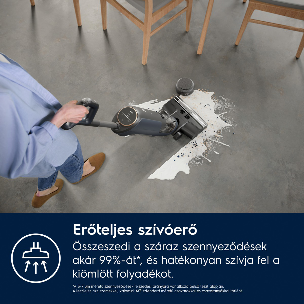Egy személy egy sötétszürke porszívót használ egy szürke padlón. A porszívó egy fehér kiömlést tisztít. Fa székek a háttérben.