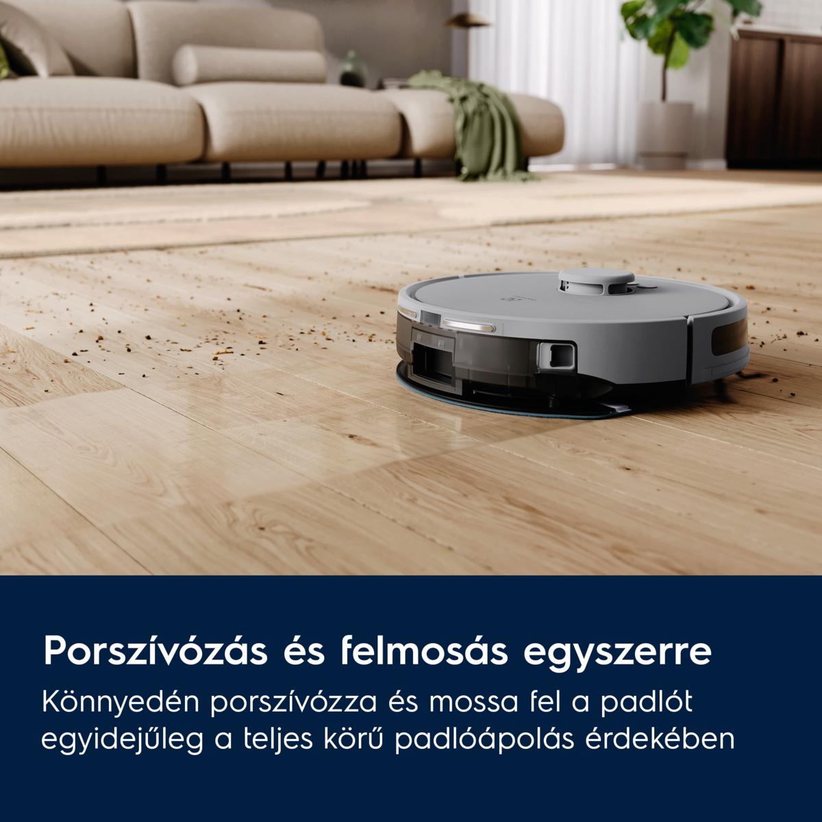 Egy szürke robotporszívó tisztít egy fa padlót. Egy bézs kanapé a háttérben. Szórt törmelék látható.