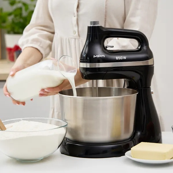 Osoba wlewa mleko ze szklanej butelki do miski miksera. Mąka, masło i czarny mikser znajdują się na stole.