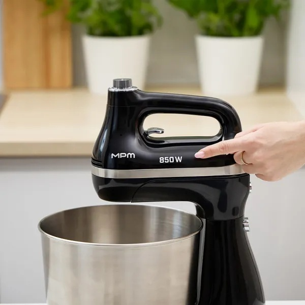 Czarny mikser stojący ze srebrną misą. Ręka wskazuje przycisk zasilania. Tło zawiera rośliny i drewnianą półkę.