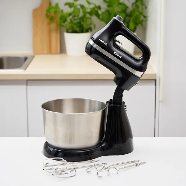 Czarny mikser stojący z misą i trzepaczkami ze stali nierdzewnej. Otoczenie kuchni z roślinami w tle, drewniana deska do krojenia.