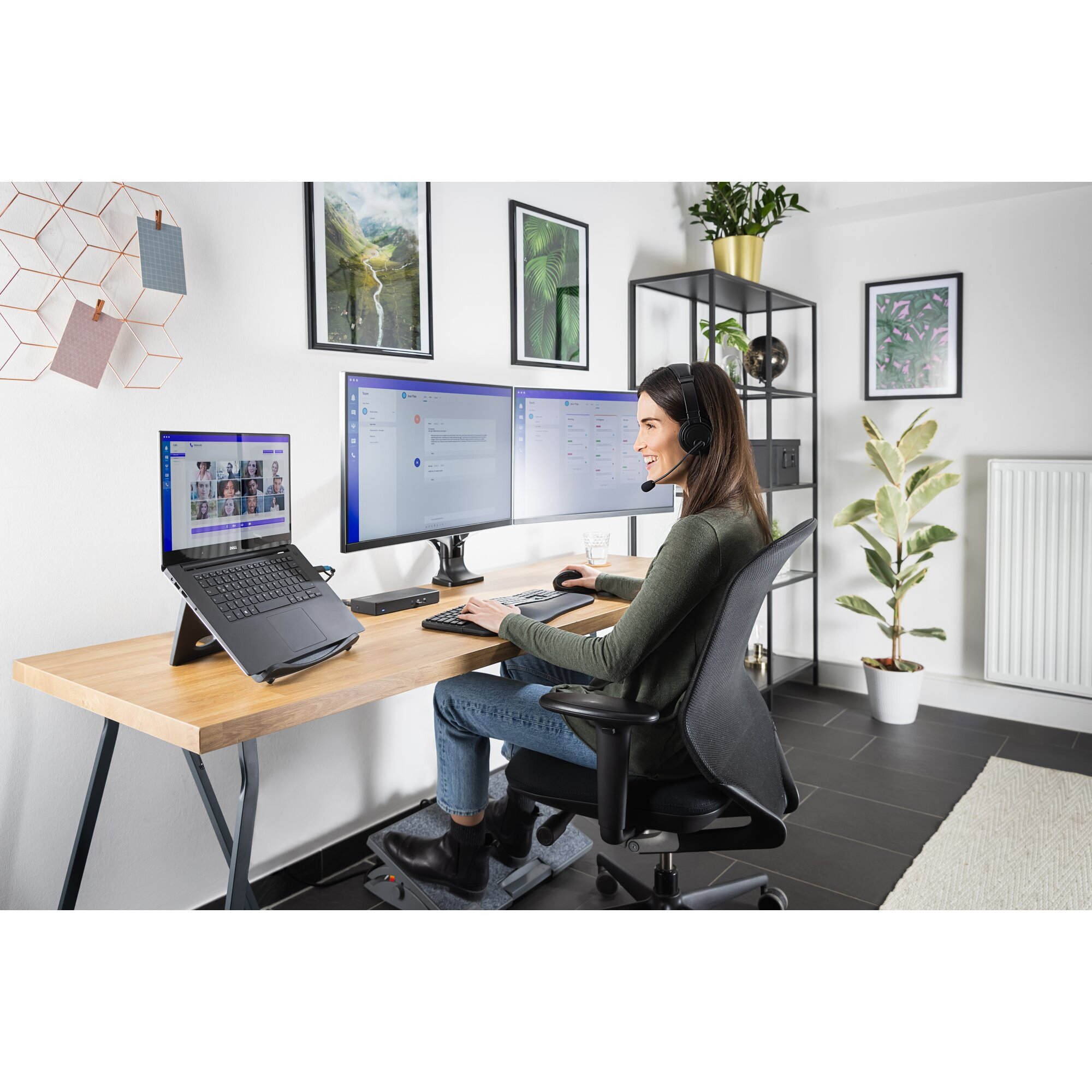 Frau arbeitet an einem Schreibtisch mit zwei Monitoren, Laptop und Headset. Sie lächelt. Pflanzen und Bilder sind im Hintergrund.