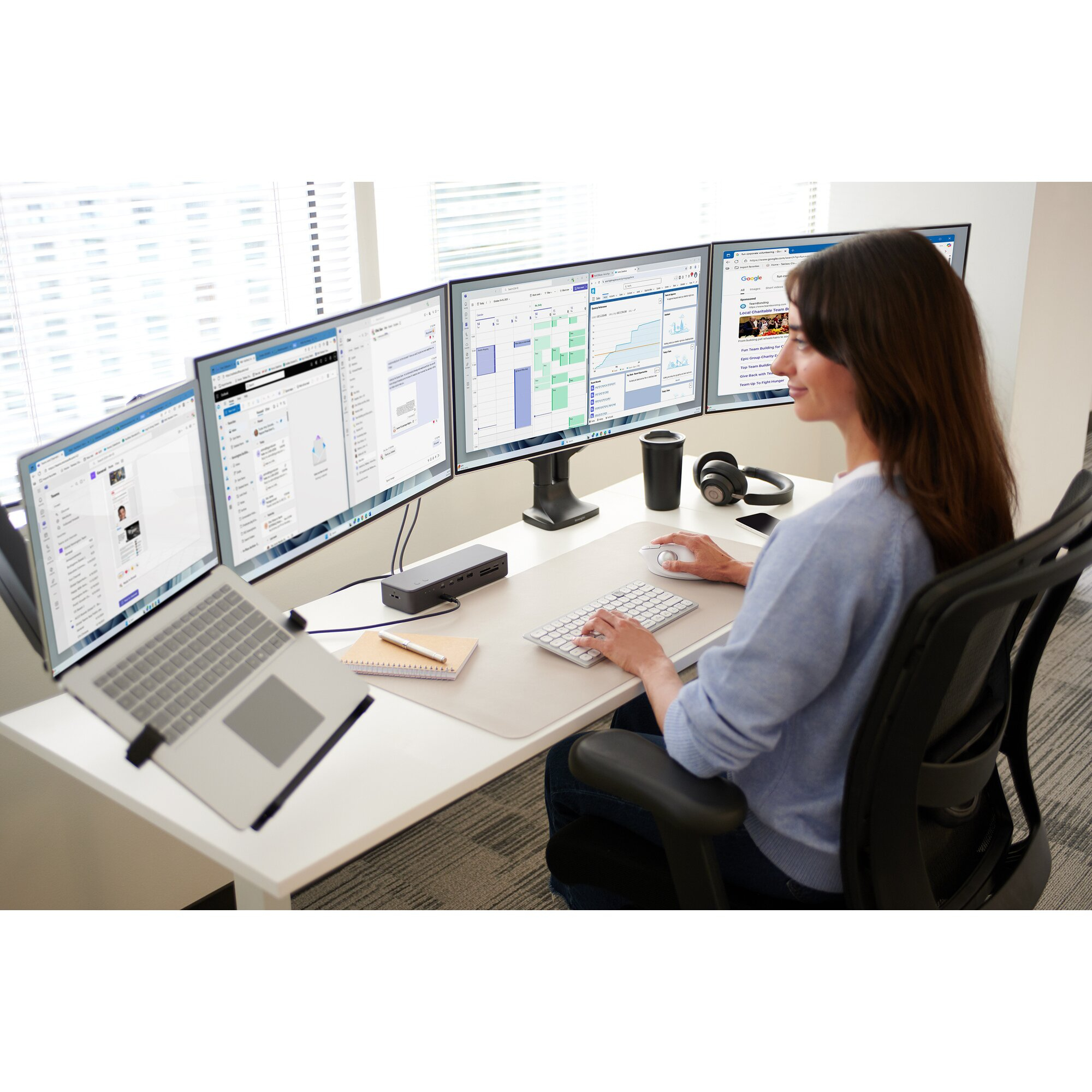 Frau an einem Schreibtisch mit mehreren Monitoren, Laptop und Zubehör. Sie arbeitet am Computer.