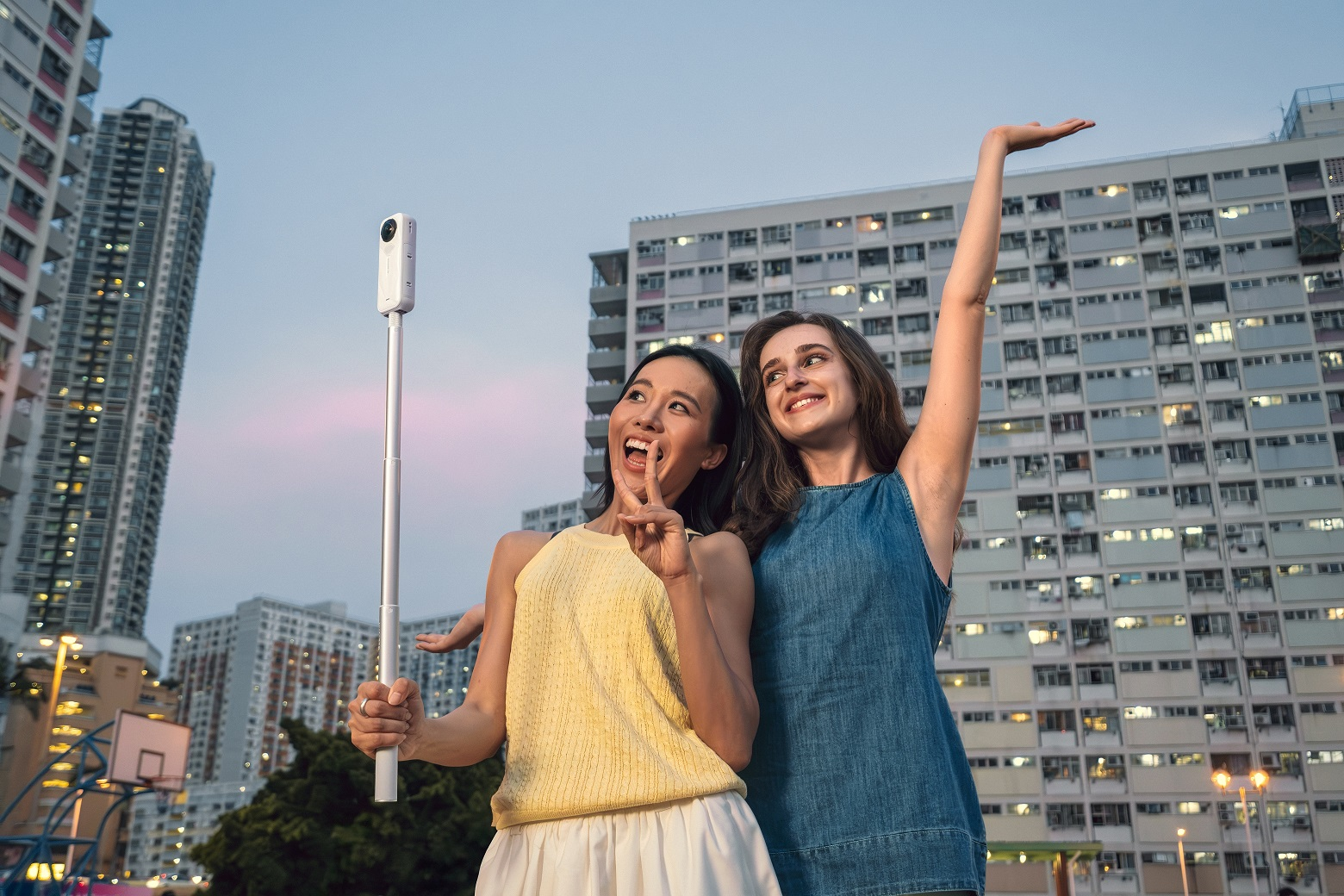 İki kadın, binaların önünde bir selfie çubuğuyla poz veriyor. Biri barış işareti yapıyor, diğeri kolunu kaldırıyor.