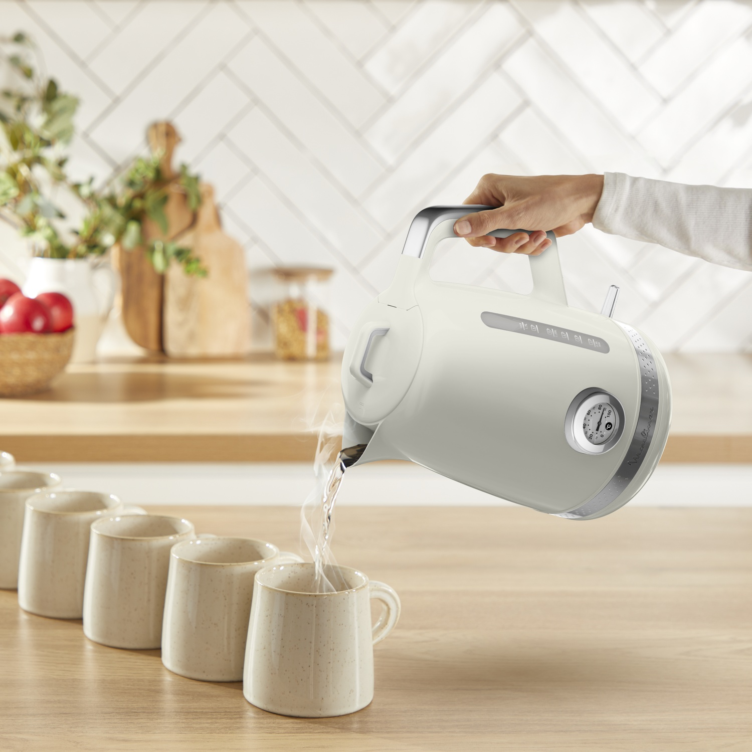 A hand pours hot water from a cream-colored kettle into mugs on a wooden table. Background has kitchen items and white tiles.