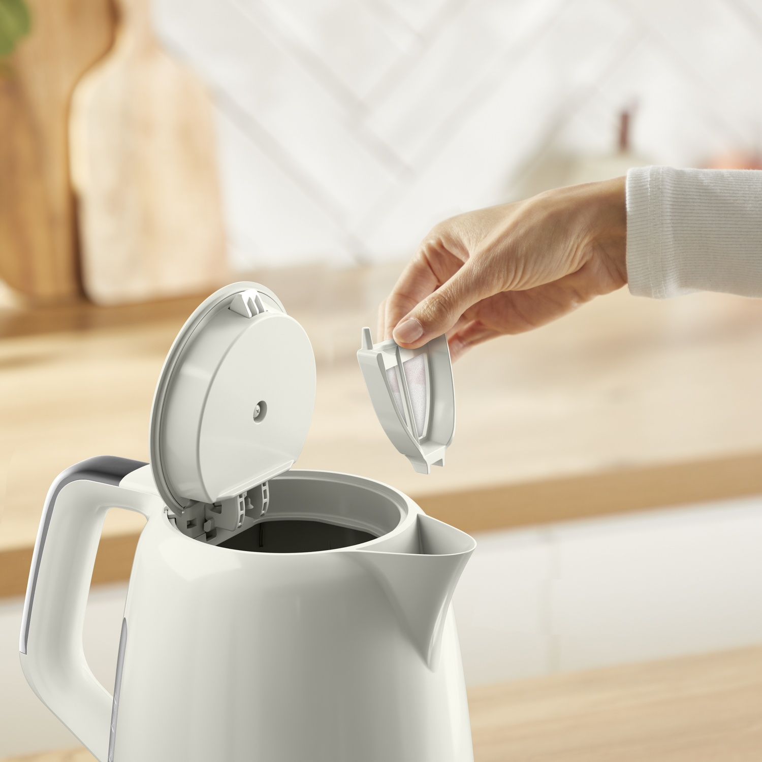 A hand lifts the lid of a white electric kettle, removing a filter. The background has a wooden surface and a tiled wall.