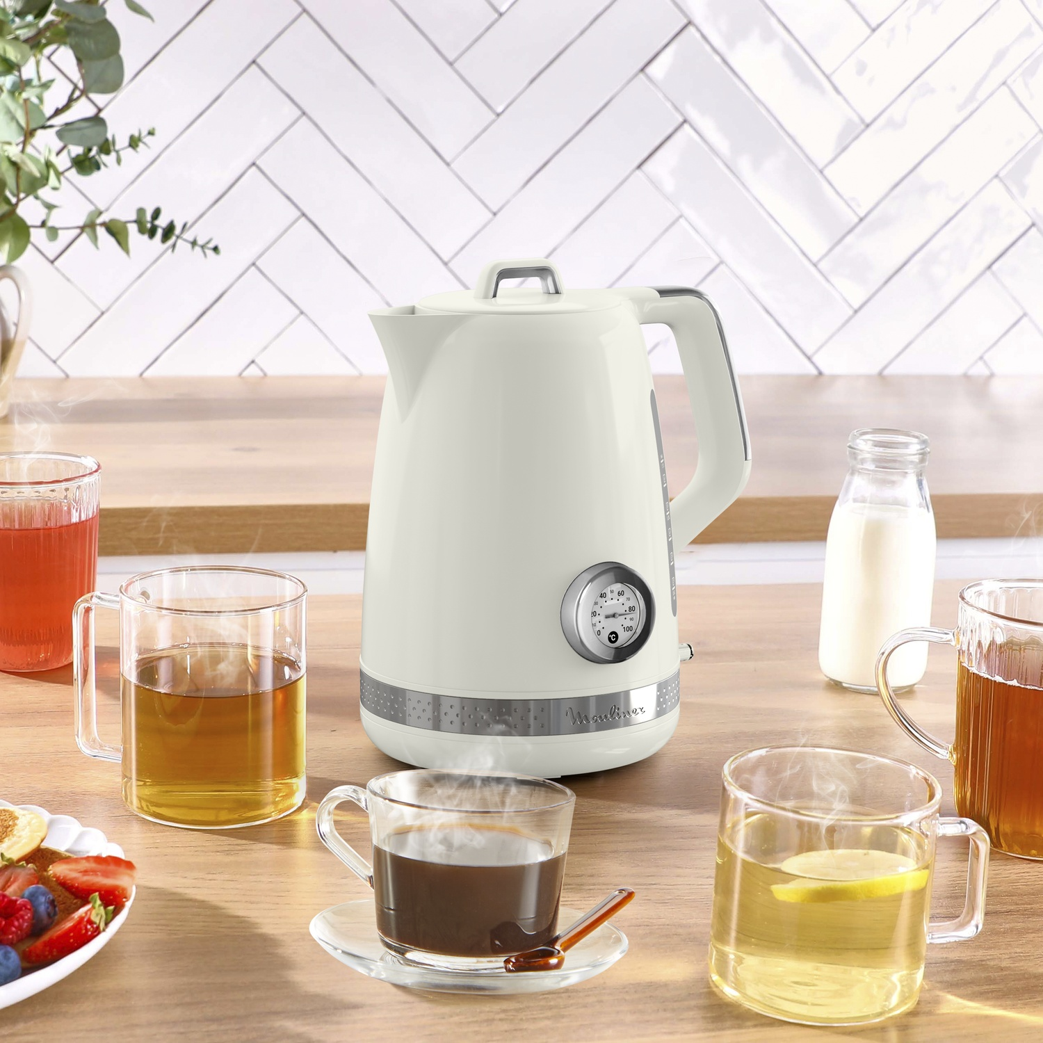 A cream kettle sits on a wood table, surrounded by glasses of tea, coffee, and milk. A plate of fruit is visible.