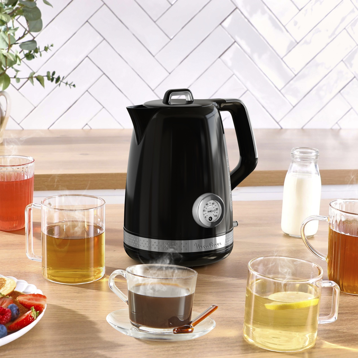 Une bouilloire noire, des tasses transparentes avec du thé, une bouteille de lait et une assiette de fruits sont sur une table en bois. Un mur carrelé blanc est en arrière-plan.