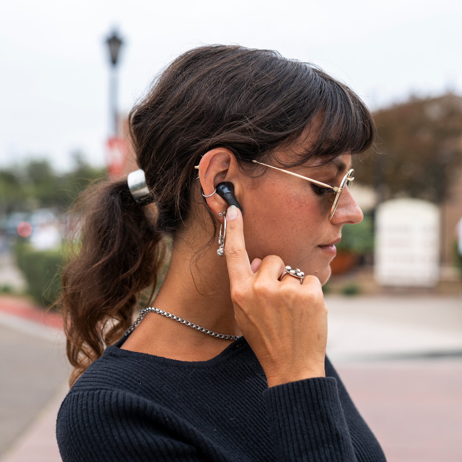 Woman wearing glasses and earbuds, touching her ear. She is wearing a black sweater, silver jewelry, and has brown hair.