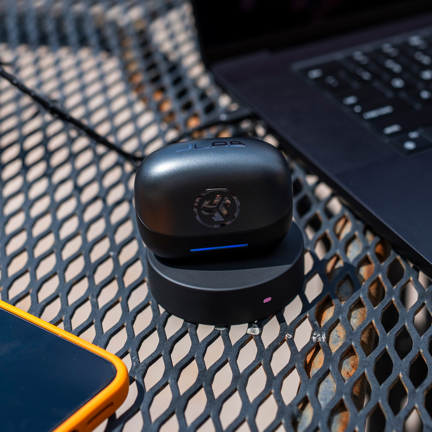 Black earbuds on a charging base, with a laptop and phone visible. The surface is a metal mesh table.