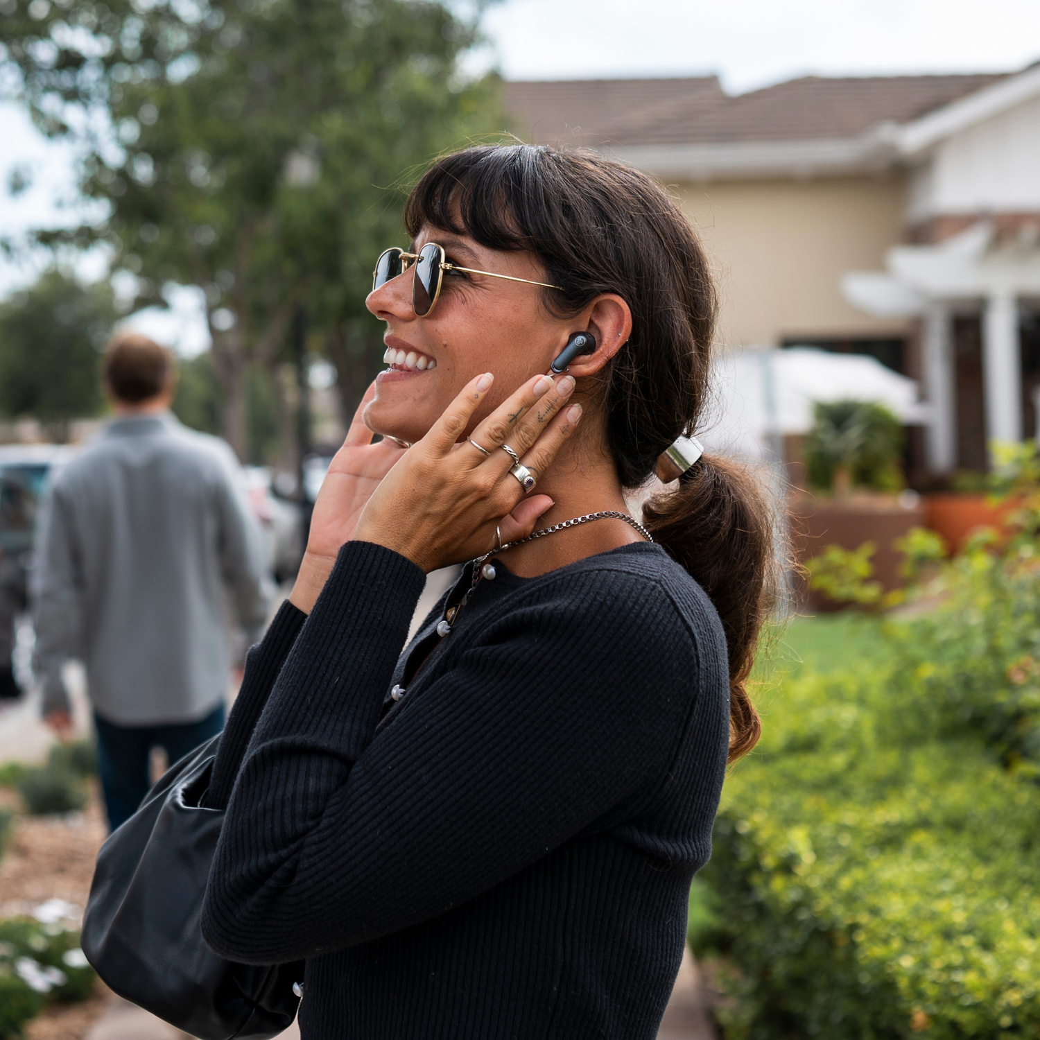 Woman wearing sunglasses and earbuds smiles outdoors. She holds her hands near her face. A man walks in the background.