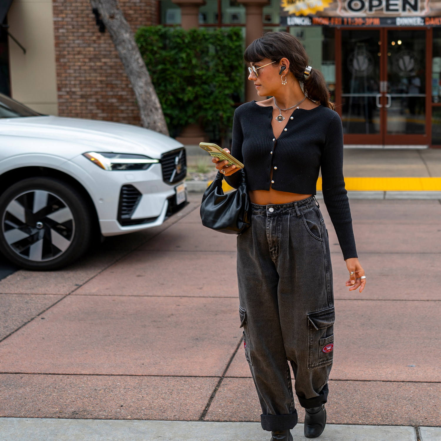 Woman walking on the sidewalk, holding phone and bag. Wearing black top, grey jeans. White car in the background.