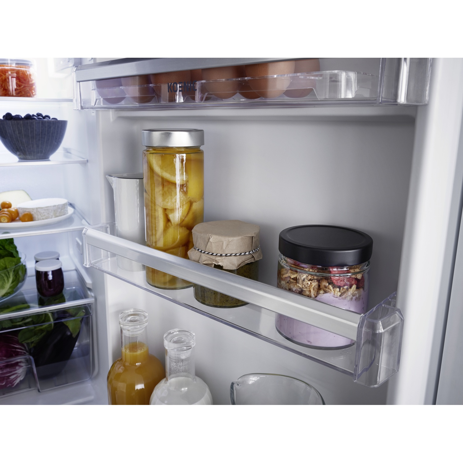 À l'intérieur d'un réfrigérateur. Des bocaux de nourriture comme des fruits et du pesto sont visibles, ainsi que des œufs, du fromage et des bouteilles de jus.
