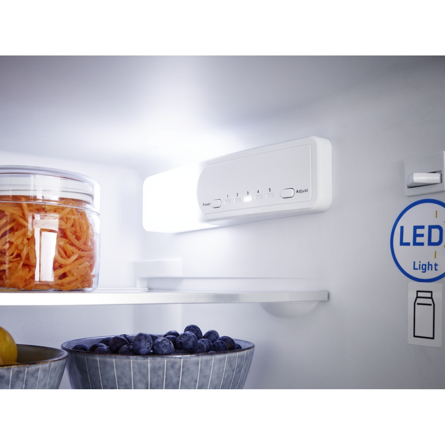 À l'intérieur d'un réfrigérateur : un bocal de carottes, un bol de myrtilles et un panneau lumineux avec des réglages. Symbole de lumière LED visible.