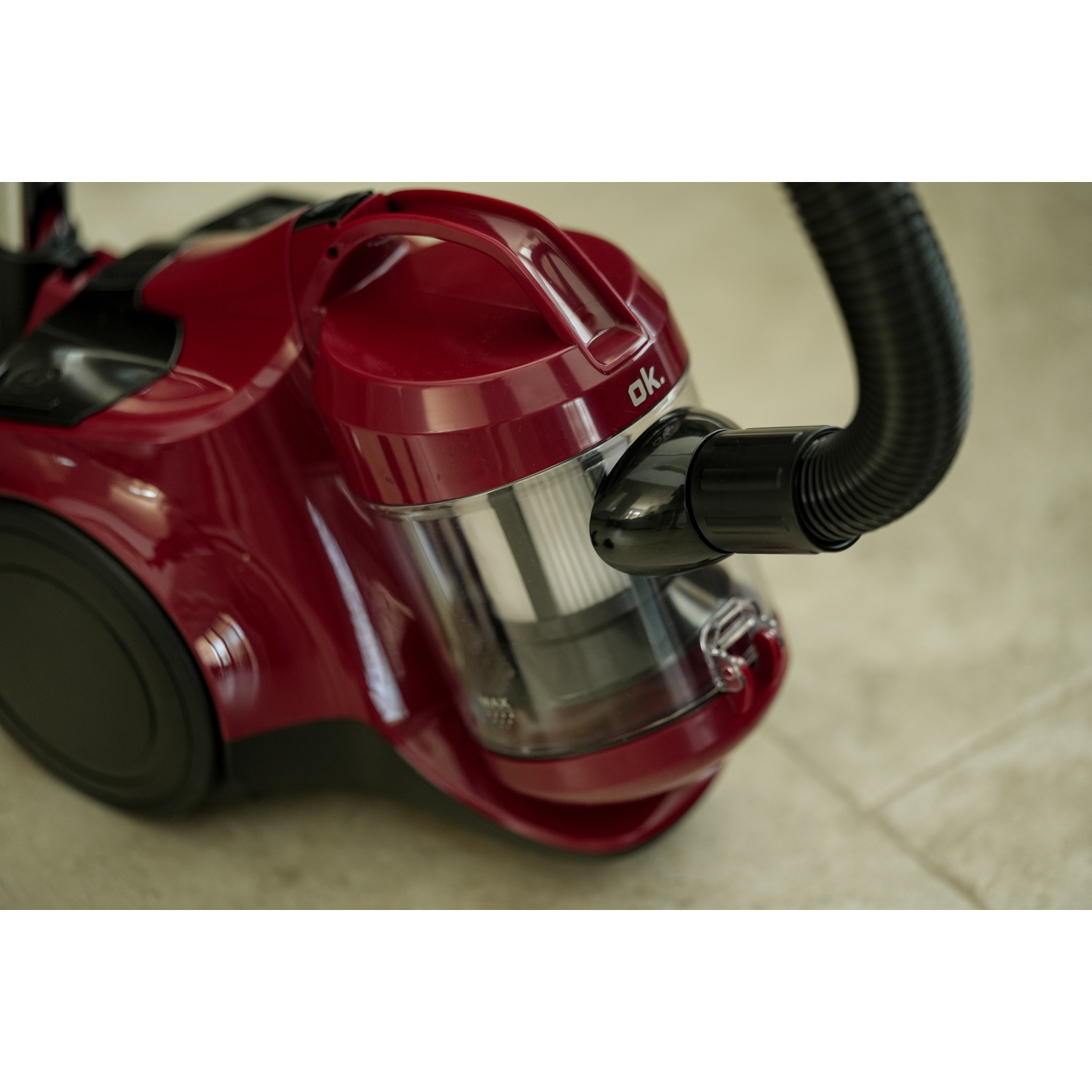 A red vacuum cleaner with a black hose. The cleaner has a clear dust container and is on a light-colored floor.